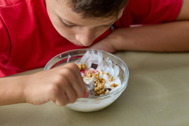 8χρονο αγόρι που τρώει μούσλι και γιαούρτι φορώντας κόκκινο μπλουζάκι, κοίτα από κοντά - Φωτογραφία, εικόνα