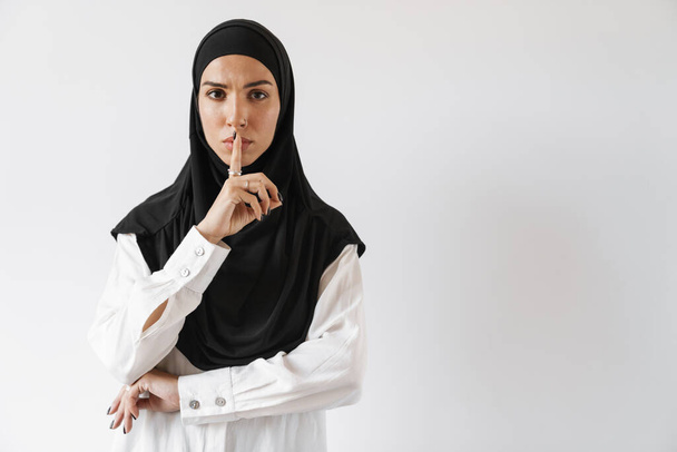 A confident muslim woman in hijab showing a silence gesture and looking at the camera in the white studio - Valokuva, kuva