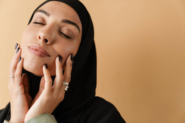 Um retrato da mulher muçulmana vestindo hijab preto e de mãos dadas perto do rosto com os olhos fechados no estúdio laranja - Foto, Imagem