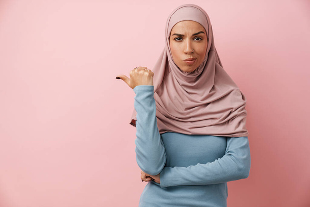 A muslim woman wearing pink hijab pointing to the copyspace and wrinkling her forehead in the pink studio - Foto, afbeelding