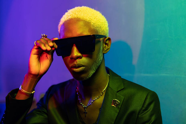 Portrait photo of a dark-skinned guy with blond hair. Dressed in a black jacket and sunglasses. With metal ornaments on the neck and arms. Purple and blue light - 写真・画像