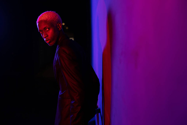 A guy with dark skin and white hair stands near the wall in a black suit. A purple light shines on his face - Foto, afbeelding