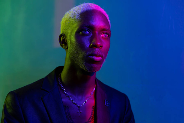 dark-skinned guy with a white face looking at a purple light. Dressed in a black jacket and silver jewelry around his neck - Fotografie, Obrázek