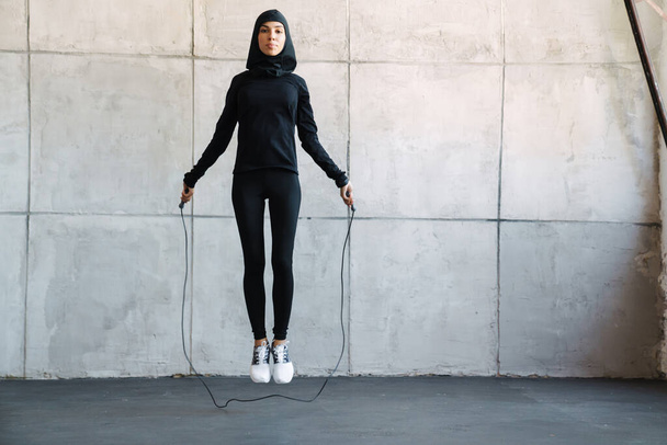 Jonge moslim sportvrouw in hijab uit te werken met springtouw binnen - Foto, afbeelding