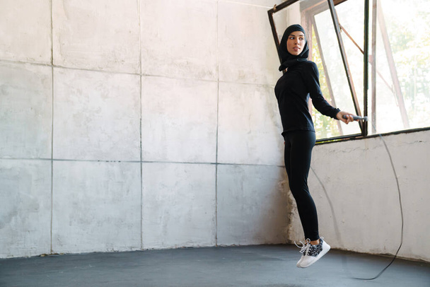 Young muslim sportswoman in hijab working out with jumping rope indoors - Valokuva, kuva