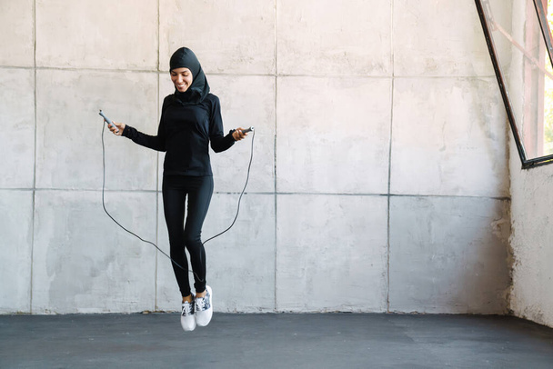 Young muslim sportswoman in hijab working out with jumping rope indoors - Foto, afbeelding