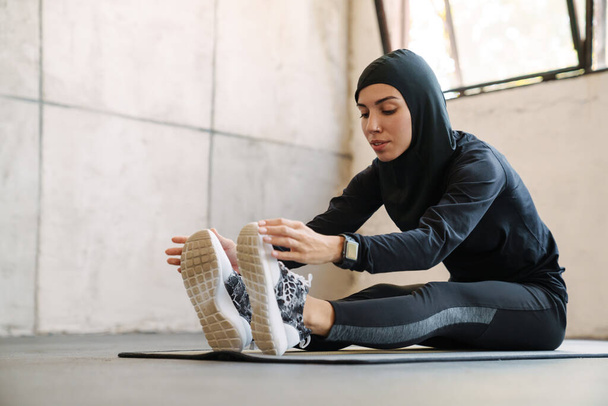 Young muslim sportswoman in hijab doing exercise while working out indoors - 写真・画像