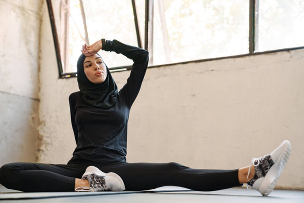 Young muslim sportswoman in hijab resting while working out indoors - Фото, зображення