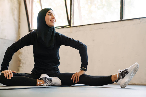 Young muslim sportswoman in hijab smiling while working out indoors - Foto, Bild