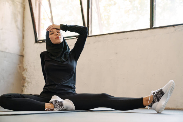 Young muslim sportswoman in hijab resting while working out indoors - Фото, изображение