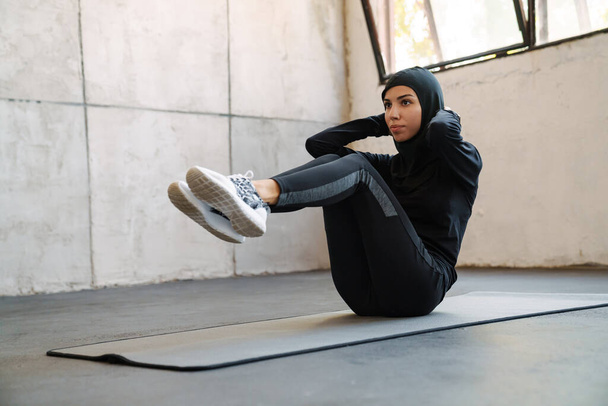 Young muslim sportswoman in hijab doing exercise while working out indoors - Photo, Image