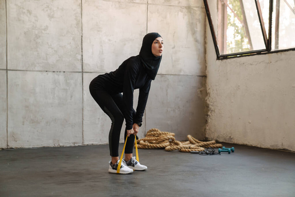 Young muslim sportswoman in hijab doing exercise with expander stretch tape indoors - Photo, Image