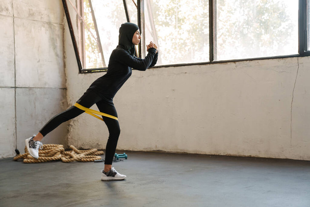 Young muslim sportswoman in hijab doing exercise with expander stretch tape indoors - Fotoğraf, Görsel