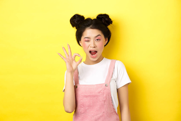 Alright. Confident asian female model with bright makeup and summer clothes, showing okay sign, winking and say yes, agree and approve, yellow background - Photo, Image