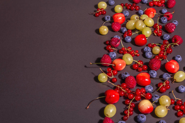 Assorted ripe berries, scattered on a black stone background. Fresh raspberries, blueberries, grape, sweet cherries, and red currants. A modern hard light, dark shadow, top view - Фото, зображення