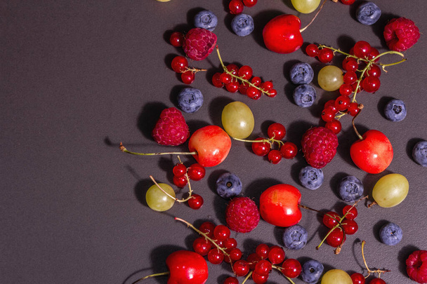 Assorted ripe berries, scattered on a black stone background. Fresh raspberries, blueberries, grape, sweet cherries, and red currants. A modern hard light, dark shadow, top view - Foto, Imagen