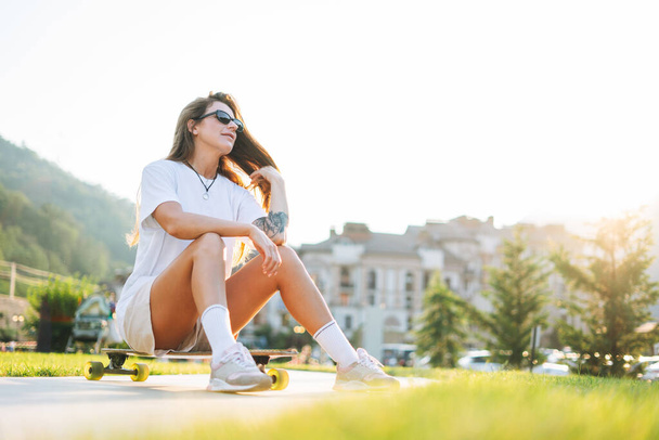 Sottile giovane donna con lunghi capelli biondi in abiti sportivi leggeri seduta con longboard nello skatepark all'aperto al tramonto - Foto, immagini