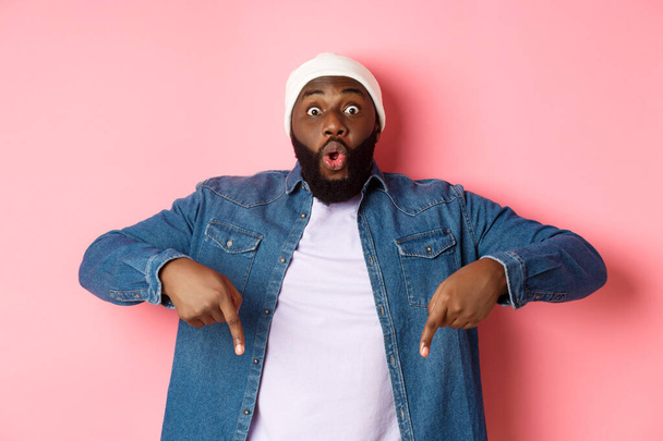 Niesamowity i pod wrażeniem afro-amerykański facet pokazujący reklamę, wpatrujący się w aparat szczęśliwy i wskazujący palcem w dół, różowe tło - Zdjęcie, obraz