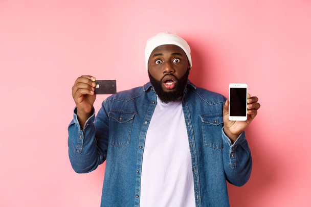 Zakupy online. Zszokowany i zaniepokojony Murzyn gapiący się w kamerę, pokazujący ekran telefonu komórkowego i kartę kredytową, stojący nad różowym tłem - Zdjęcie, obraz
