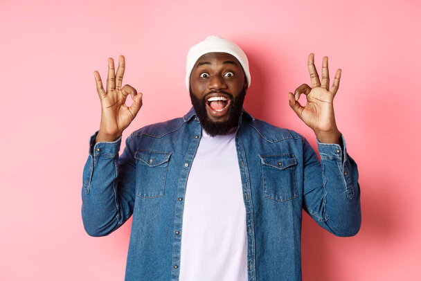Impresionado hombre negro en gorro y camisa, mostrando signos de ok y mirando a la cámara sorprendida, sonriendo feliz, alabanza gran oferta, recomendando el producto, fondo rosa - Foto, imagen