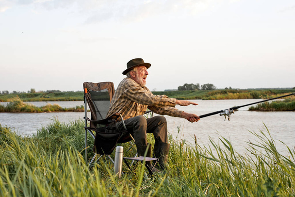 Ηλικιωμένος άνδρας που ψαρεύει στη λίμνη, ψάρεμα ως χόμπι, πλαϊνή άποψη πορτρέτο, στην ύπαιθρο - Φωτογραφία, εικόνα
