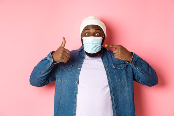 Coronavirus, estilo de vida y concepto de pandemia global. Joven hombre afroamericano apuntando a la máscara facial y mostrando los pulgares hacia arriba, protegerse de covid, fondo rosa - Foto, imagen