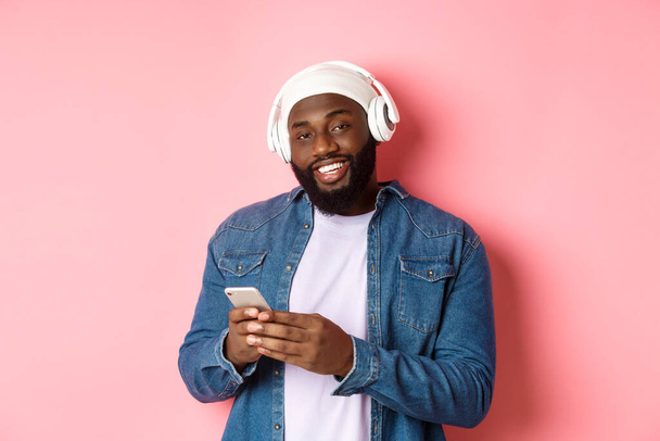 Όμορφος hipster τύπος με ακουστικά χαμογελά ικανοποιημένοι στην κάμερα, ακούγοντας μουσική στα ακουστικά, χρησιμοποιώντας το κινητό app, στέκεται πάνω από ροζ φόντο - Φωτογραφία, εικόνα