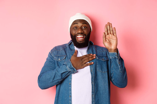 Изображение улыбающегося африканско-американского человека, честного, говорящего правду, руки на сердце и в воздухе, дающего обещания или ругающегося, стоящего на розовом фоне - Фото, изображение