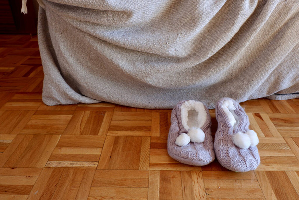 Zapatillas de casa de punto zapatos en piso de madera cerca de sofá cubierto con manta. Conceptual para momentos de la vida real - Foto, imagen