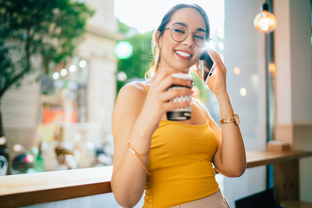 Funny young woman in optical eyewear enjoying friendly smartphone conversation in roaming, cheerful hipster girl with coffee to go smiling while connecting to 4g internet for making international call - Foto, Bild