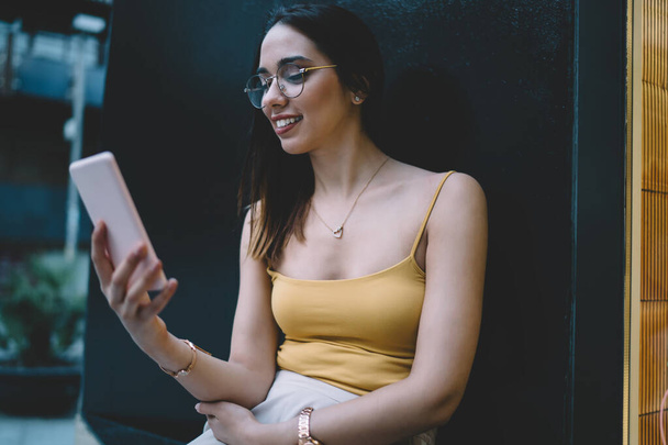 Happy Caucasian woman sending text messages and reading information on mobile phone resting outdoors, smiling female in classic eyewear watching positive video online on smartphone technology - Photo, image