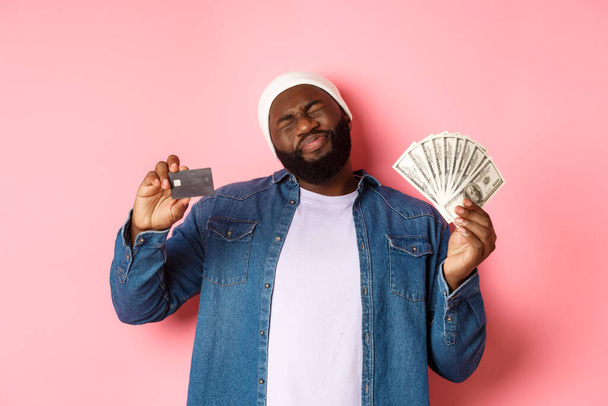 Shopping concept. Sad and whining Black guy showing credit card and dollars, grimacing reluctant, standing over pink background - Photo, Image