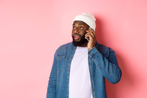 Hipster Czarny mężczyzna rozmawia przez telefon, patrzy w lewo i prowadzi mobilną rozmowę, stoi nad różowym tle - Zdjęcie, obraz