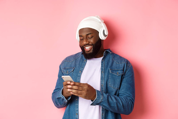 Charmant homme noir élégant écoutant de la musique dans les écouteurs et écrivant un message sur smartphone, souriant heureux, debout sur fond rose - Photo, image