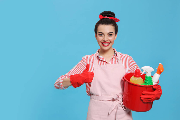 Young housewife holding bucket with cleaning supplies on light blue background, space for text - Foto, afbeelding