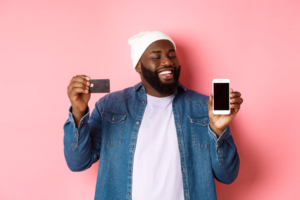 Online winkelen. Tevreden Zwarte man knikt in goedkeuring, glimlacht en kijkt naar telefoon, toont credit card en smartphone scherm, staan over roze achtergrond - Foto, afbeelding
