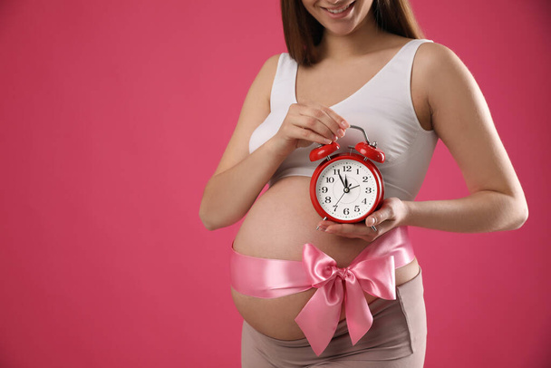 Young pregnant woman with alarm clock and bow on pink background, closeup. Time to give birth - Foto, imagen