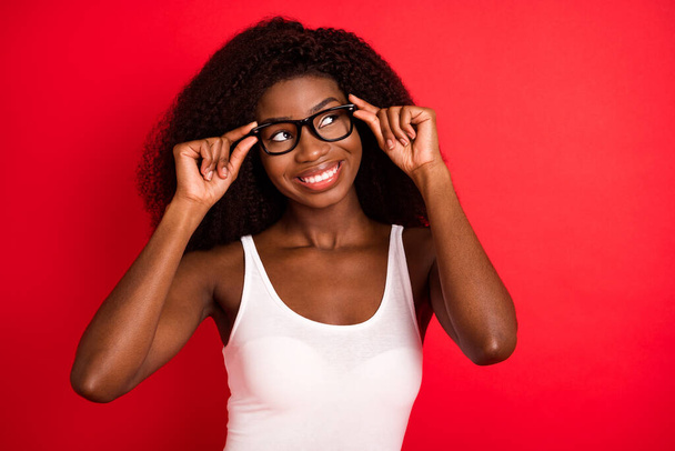 Foto de joven atractiva afro chica feliz sonrisa positiva manos tacto gafas mirada espacio vacío aislado sobre fondo de color rojo - Foto, Imagen