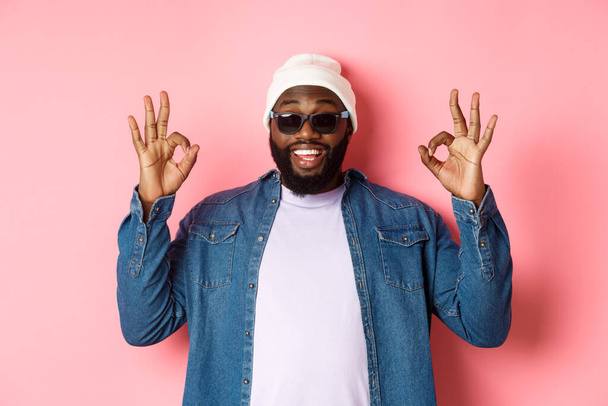 Image of cool african-american hipster man showing okay signs in approval, like something awesome, wearing sunglasses and beanie, pink background - Photo, Image