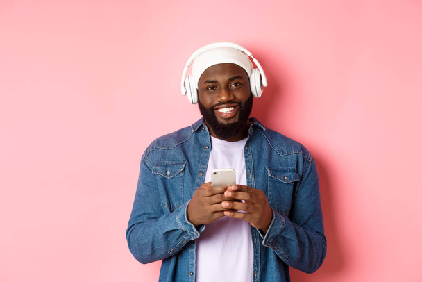 Image d'un beau Noir dans un casque, écoutant de la musique et utilisant un téléphone portable, souriant à la caméra, fond rose - Photo, image