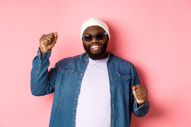 Happy Black man in beanie an sunglasses rejoicing, dancing with happy face, standing over pink background - Photo, Image