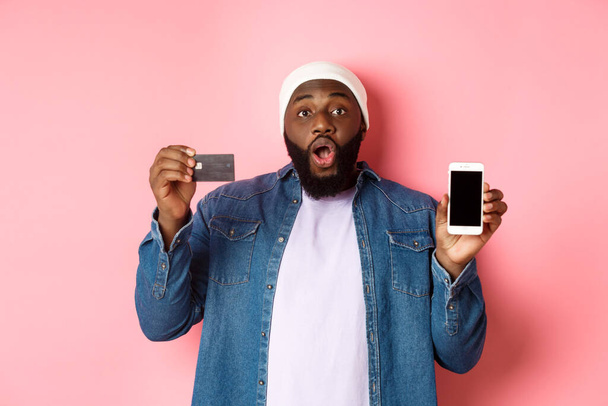 Online αγορές. Ενθουσιασμένος Μαύρος άνδρας που δείχνει πιστωτική κάρτα και οθόνη του κινητού τηλεφώνου, στέκεται πάνω από ροζ φόντο έκπληκτος - Φωτογραφία, εικόνα
