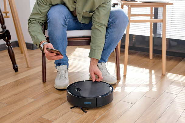 Young cropped male using automatic smart robot vacuum cleaner working on floor - Foto, Imagen