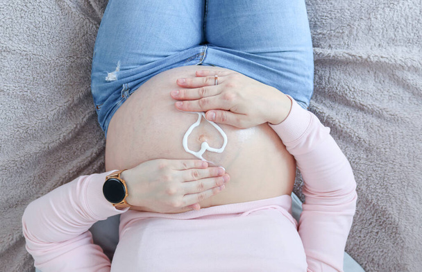 Hamile bir kadın, göbeğini nemlendiriciden yapılmış bir kalple tutar.. - Fotoğraf, Görsel