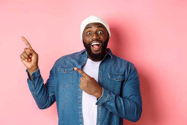 Εντυπωσιασμένος μαύρος τύπος που δείχνει φοβερή προσφορά promo, κοιτάζοντας κάμερα ενθουσιασμένοι και χαμογελώντας, δείχνοντας τα δάχτυλα στην πάνω αριστερή γωνία πανό, ροζ φόντο - Φωτογραφία, εικόνα
