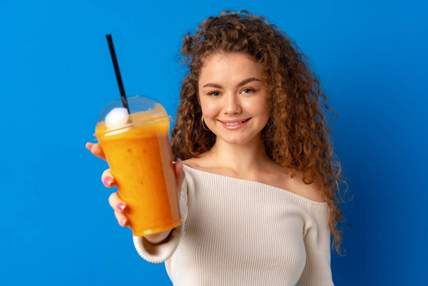 Beautiful curly-haired woman drinking orange juice against blue background - Zdjęcie, obraz