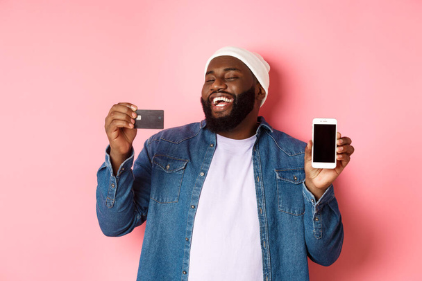Compras en línea. Feliz hombre afroamericano en gorro riendo, mostrando la tarjeta de crédito y la pantalla del teléfono móvil, de pie sobre el fondo rosa - Foto, imagen