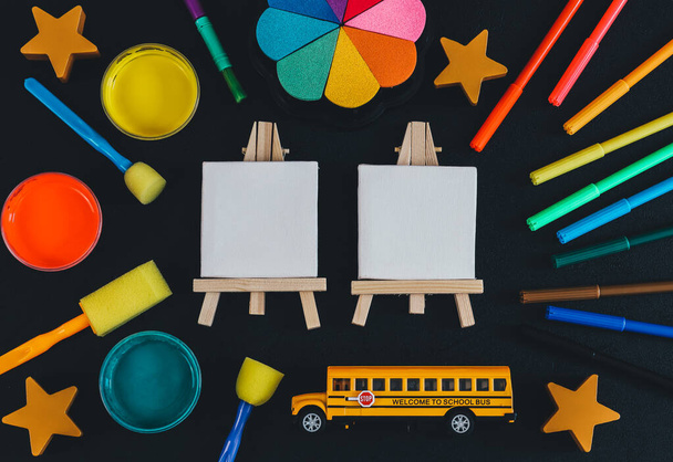 Egy iskolabusz festőállványokkal, festékekkel, filctollakkal és egy jegyzettömbbel fekete alapon fekszik, középen szöveges háttérrel, felülnézeti közeli képekkel.. - Fotó, kép