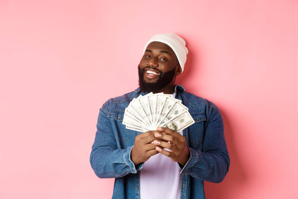 Задоволений багатий афро-американський чоловік показує гроші, посміхаючись задоволений і показуючи з доходом, стоїть з доларами на рожевому тлі
 - Фото, зображення