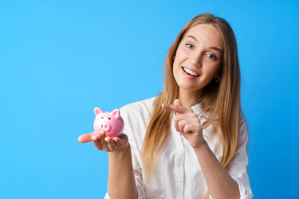 Beautiful smiling young woman with piggy bank on blue background - Photo, image